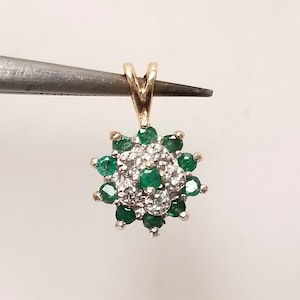 May include: A gold pendant featuring a central cluster of small, clear, round-cut stones surrounded by a halo of ten vibrant green, round-cut stones. The pendant has a triangular gold bail at the top, designed for attachment to a necklace chain.