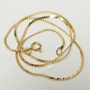 May include: A gold chain necklace featuring a flat, wavy, serpentine link design. The necklace includes a circular spring ring clasp and a small jump ring at the end, showcasing a polished, reflective metallic finish throughout the entire piece.