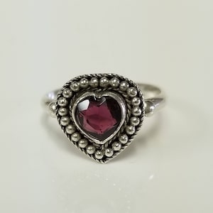 May include: A silver ring featuring a faceted heart-shaped dark red gemstone at the center. The stone is surrounded by a decorative border of small silver beads and a twisted rope-like metal frame, set on a smooth, polished silver band.