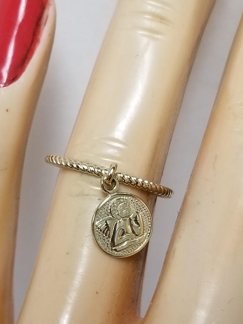 May include: A gold ring with a twisted band and a small charm of an angel with wings. The charm is round and has a detailed design.