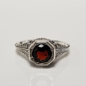 May include: A silver ring featuring a round, faceted deep red garnet gemstone set within an octagonal frame. The band and setting display intricate, openwork metal filigree detailing that extends along the sides of the ring shank.