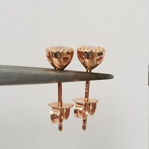 May include: A pair of rose gold stud earrings with a round, clear stone in the center. The earrings have a detailed, textured design around the stone.