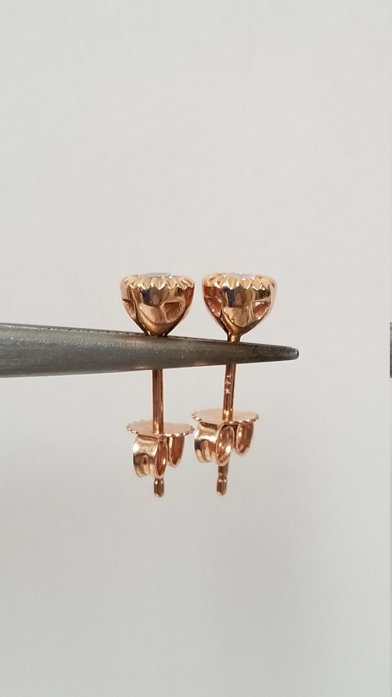 May include: A pair of rose gold stud earrings with a round, clear stone in the center. The earrings have a detailed, textured design around the stone.