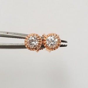 May include: A pair of rose gold stud earrings with a round, clear diamond in each setting. The diamonds are surrounded by a delicate, textured halo.