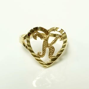 May include: A gold-colored ring featuring a heart-shaped design with the letter "K" in the center. The ring has a textured, ridged border around the heart. The ring is set against a white background.