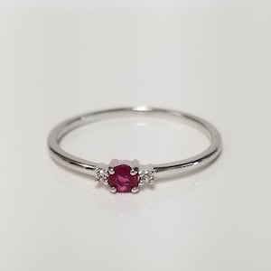May include: A delicate silver ring with a single red ruby gemstone and two smaller diamonds on either side.