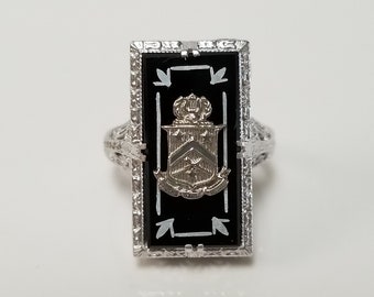 May include: A silver ring with a black onyx stone set in a rectangular frame. The stone features a gold crest with a crown and other details.