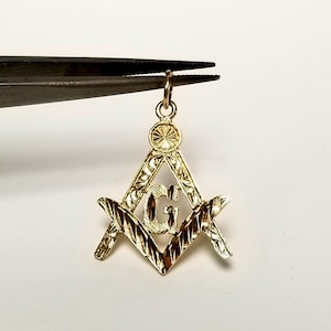 May include: A gold-colored pendant featuring the Masonic Square and Compasses symbol. The pendant has a detailed design with the letter "G" in the center. The pendant is held by a pair of tweezers against a white background.