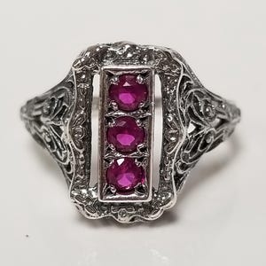 May include: A silver-toned ring featuring an elongated rectangular center setting with three round, vibrant pinkish-red stones arranged vertically. The band and the frame surrounding the stones display intricate, openwork metal filigree detailing.