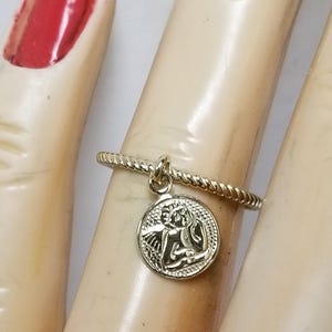 May include: Gold ring with a twisted band and a charm of an angel with wings. The charm is round and has a detailed design.