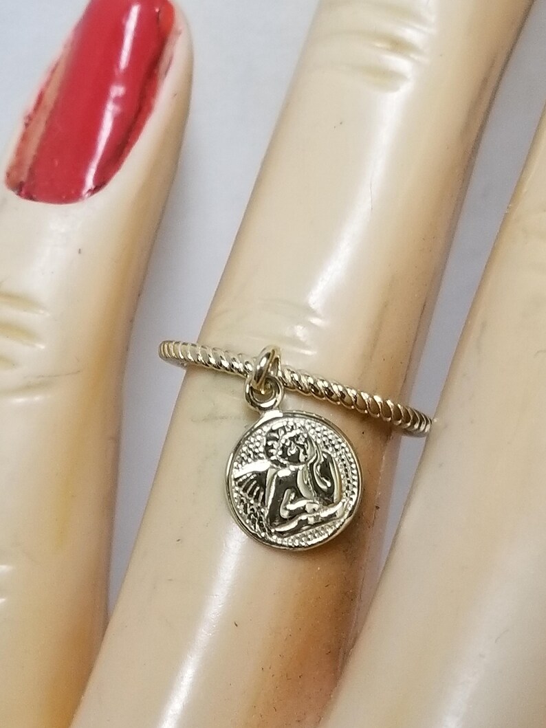 May include: Gold ring with a twisted band and a charm of an angel with wings. The charm is round and has a detailed design.