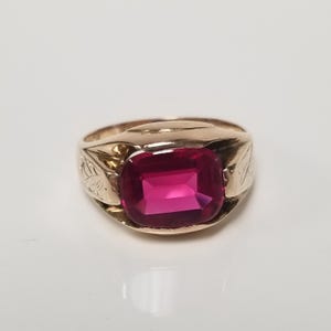 May include: A gold ring featuring a large, rectangular, faceted deep pink gemstone set in the centre. The band of the ring displays an engraved leaf pattern on both sides, extending from the central setting towards the back of the band.