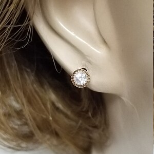 May include: A close-up of a gold earring with a clear round stone in the center. The earring is set in a small, round, gold frame with a textured edge.