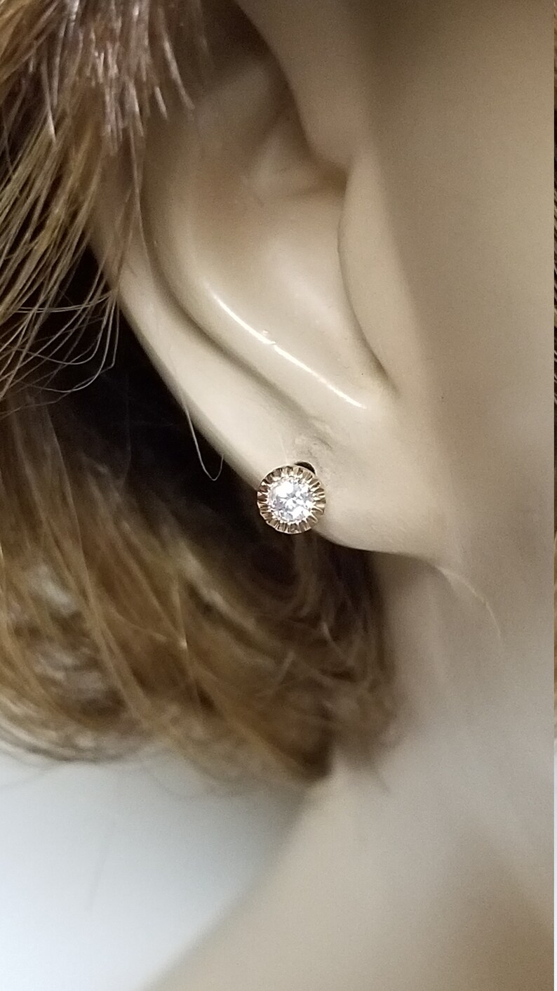 May include: A close-up of a gold earring with a clear round stone in the center. The earring is set in a small, round, gold frame with a textured edge.