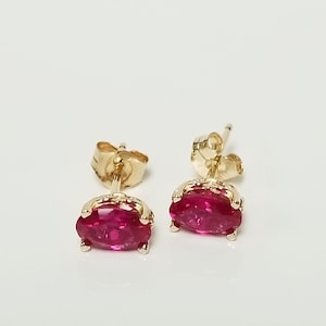 May include: A pair of gold stud earrings with oval-shaped, vibrant red gemstones. The earrings have a classic design with a post and butterfly backing. The gemstones are set in a gold bezel setting, adding a touch of elegance.