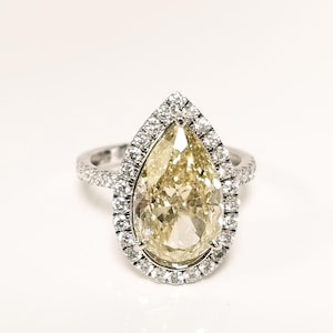 May include: A pear-shaped diamond ring with a halo of smaller diamonds. The large, central diamond has a yellow hue, and the band is encrusted with additional diamonds. The ring is set against a white background.