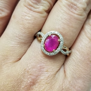May include: A gold ring featuring a large oval-cut ruby gemstone surrounded by a halo of small, clear diamonds. The ring's band has a twisted design. The ruby is a vibrant pinkish-red color.