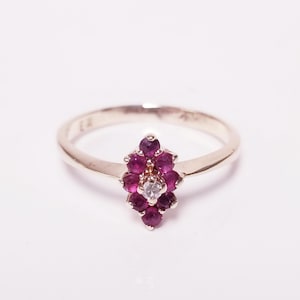 May include: A gold-toned ring featuring a cluster of small, deep red gemstones arranged in a floral pattern. A single, clear gemstone is centered within the cluster. The ring is set against a white background.