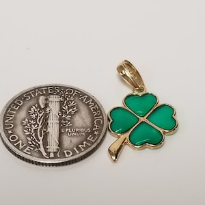 May include: A gold-colored pendant in the shape of a four-leaf clover with green enamel filling. The pendant is next to a U.S. dime coin for size comparison. The clover has a gold outline and a loop for a chain.