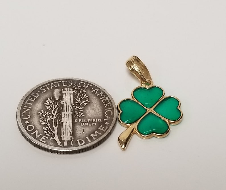 May include: A gold-colored pendant in the shape of a four-leaf clover with green enamel filling. The pendant is next to a U.S. dime coin for size comparison. The clover has a gold outline and a loop for a chain.