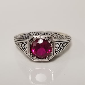 May include: A silver ring with an ornate filigree design and a large, faceted, ruby-red gemstone.