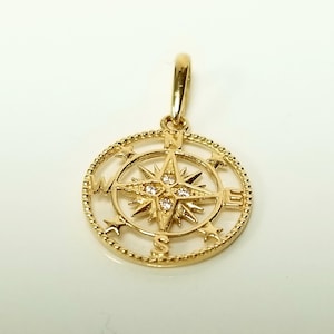 May include: A gold-colored pendant in the shape of a compass. The compass rose is set within a circular frame with small stars and a beaded border. The center of the compass features four small, clear gemstones.