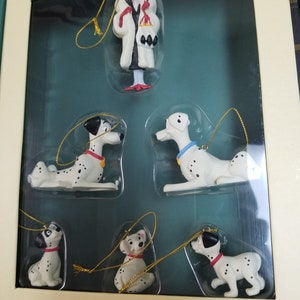 May include: A set of seven Christmas ornaments featuring characters from the Disney movie 101 Dalmatians. The ornaments include Cruella De Vil, a Dalmatian puppy, and a Dalmatian adult dog. The ornaments are packaged in a clear plastic box with a white background.