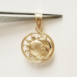 May include: A gold-colored pendant featuring a sun and moon design. The sun has a face with rays extending outward, and the moon is crescent-shaped. The pendant is held by a pair of tweezers against a white background.