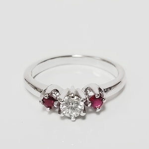 May include: A silver-toned ring featuring a central round-cut clear gemstone flanked by two smaller round-cut red gemstones. The stones are held in place by metal prongs on a smooth, polished band that forms a continuous circular shape.