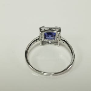 Op de afbeelding: Een zilveren ring met een vierkante blauwe edelsteen gezet in een zilveren frame. De ring ligt op een witte achtergrond.