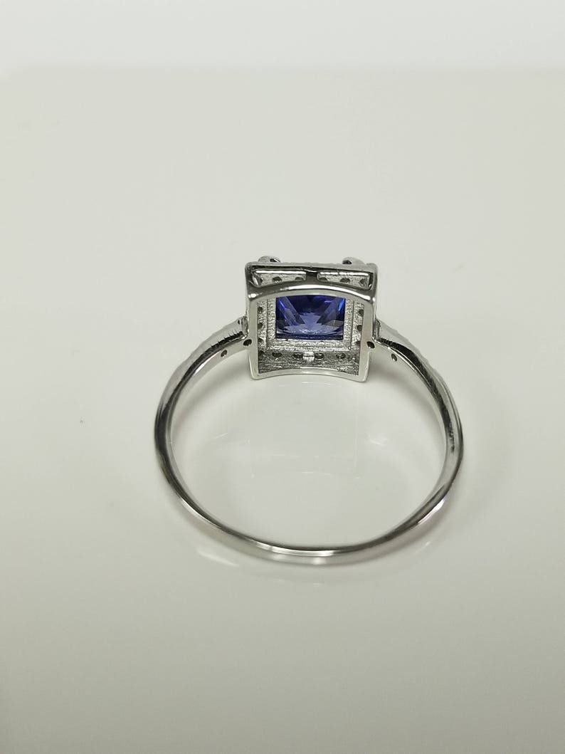 Op de afbeelding: Een zilveren ring met een vierkante blauwe edelsteen gezet in een zilveren frame. De ring ligt op een witte achtergrond.