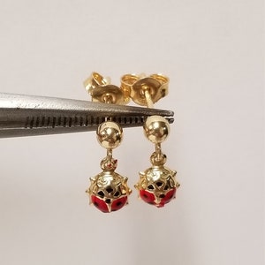 May include: A pair of gold ladybug earrings with red and black enamel. The earrings are dangle style with a small gold ball at the top.
