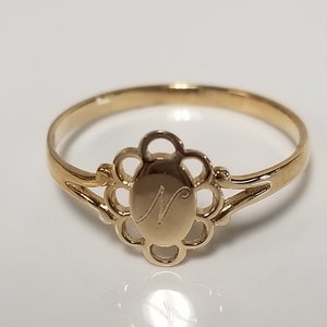 May include: A gold ring with a delicate floral design and an oval-shaped centre featuring the letter "N" engraved inside.