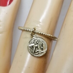 May include: A gold ring with a twisted band and a small oval charm featuring an angel with wings. The charm hangs from the ring.