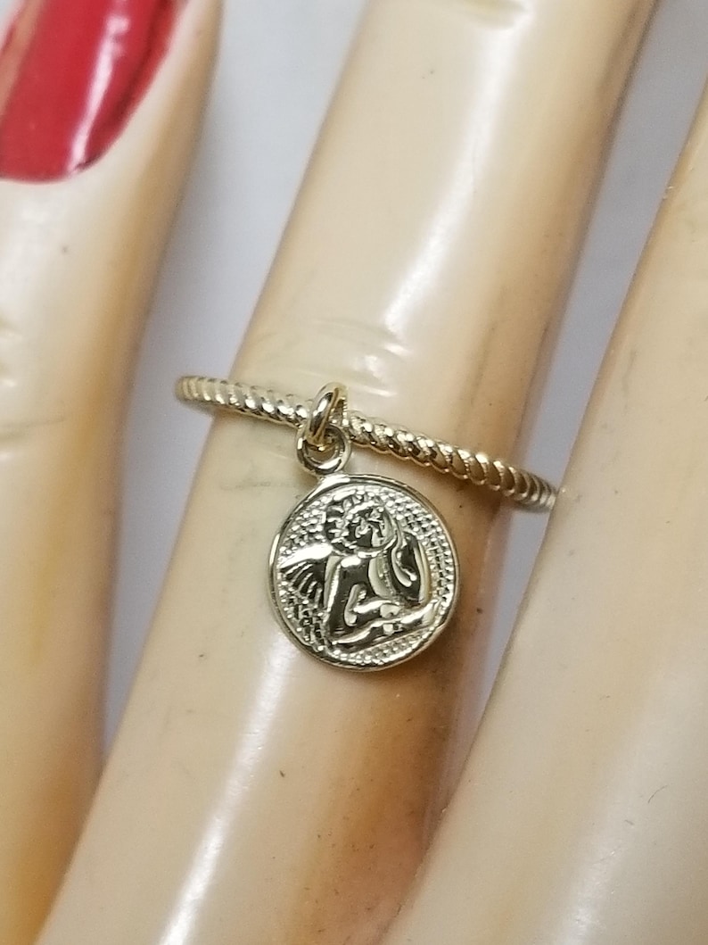 May include: A gold ring with a twisted band and a small oval charm featuring an angel with wings. The charm hangs from the ring.
