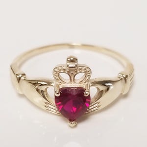 May include: A gold-colored Claddagh ring with a heart-shaped, deep red gemstone. The ring features a crown above the heart and clasped hands on either side, symbolizing love, loyalty, and friendship. The ring is a classic piece of jewelry.