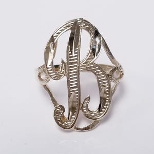 May include: Silver ring with an ornate 'B' design. The ring has a polished finish with etched details. The ring is set against a white background. A piece of jewellery.
