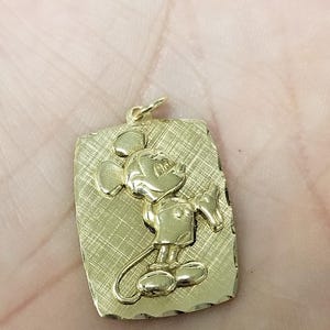 K&ouml;nnte beinhalten: Goldfarbener Anh&auml;nger mit einem erhabenen Bild von Mickey Mouse auf einem rechteckigen, strukturierten Hintergrund. Der Anh&auml;nger hat oben eine kleine Schlaufe f&uuml;r eine Kette. Das Design ist ein klassischer Zeichentrickcharakter.