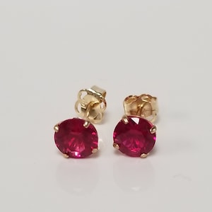May include: A pair of gold stud earrings with vibrant red gemstones. The earrings are round and set in a gold bezel. The earring backs are also gold. The earrings are displayed on a white surface.