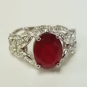 May include: A silver ring with a large, oval-shaped red gemstone set in a delicate, intricate design. The ring is adorned with smaller, clear gemstones.