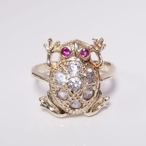 May include: A silver-toned ring featuring a frog-shaped centerpiece. The frog has two small, round, vibrant pink gemstone eyes and a back adorned with seven clear, round, faceted white stones arranged in a floral-like cluster pattern.