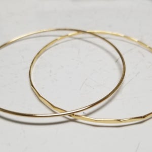 May include: Two delicate gold bangles, each with a hammered texture, are displayed on a white surface. The thin, circular bracelets overlap slightly, showcasing their simple yet elegant design. The warm gold colour contrasts against the neutral background.