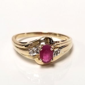 May include: A gold ring featuring a central oval-cut red ruby held in a four-prong setting. The band is crafted from polished yellow gold and includes two small, clear, round-cut accent stones set on either side of the ruby in white metal.