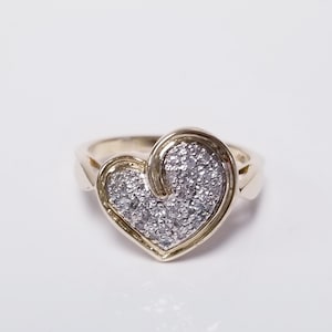 May include: A gold-toned ring featuring a heart-shaped design. The heart is filled with small, sparkling stones, creating a shimmering effect. The ring's band has a smooth, polished finish.