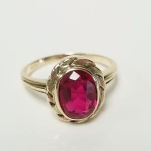 Puede incluir: Anillo dorado con una gema roja ovalada y vibrante. La gema está engastada en un bisel decorativo con un diseño trenzado. La banda del anillo es lisa y sencilla, creando un aspecto clásico y elegante.