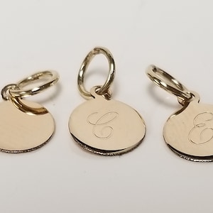 May include: Three gold-colored metal charms with a circular shape. Two charms have the letter "E" engraved on them, while the third charm is plain.