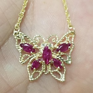 May include: A gold-toned necklace featuring a butterfly-shaped pendant with intricate filigree metalwork and five marquise-cut deep pink gemstones arranged to form the body and wings of the butterfly, suspended from a delicate gold-toned chain.