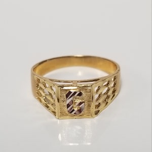 May include: A gold ring with a square design featuring the letter "G" in the centre. The ring has a delicate, intricate pattern around the edges.