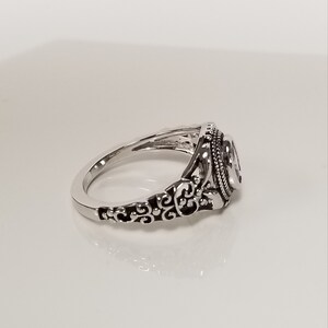 May include: A silver ring with an ornate design. The ring has a filigree pattern and a round stone in the center.