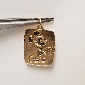 K&ouml;nnte beinhalten: Goldfarbener Anh&auml;nger mit einem erhabenen Bild von Mickey Mouse auf einer rechteckigen Platte. Der Anh&auml;nger wird von einer Pinzette gehalten. Die Platte hat eine strukturierte Oberfl&auml;che mit einem Rand.